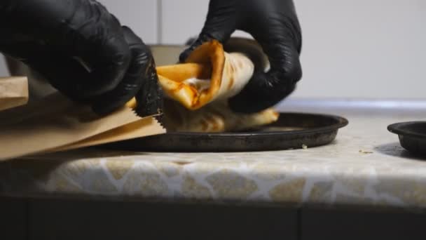 Manos masculinas de chef en guantes poniendo sabroso pan de pita con col en una bolsa de papel en la cocina. Brazos de cocinero dispuesto delicioso lavash para ser servido. Concepto de cocina de alimentos. Primer plano: cámara lenta — Vídeos de Stock