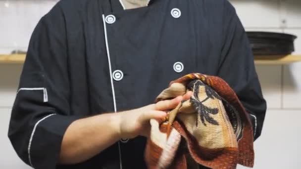 Cierre joven cocinero en uniforme de pie en el restaurante de la cocina y limpiarse las manos con una toalla después de cocinar platos. Chef masculino mirando a la cámara y sonriendo en la cafetería de la cocina. Grúa tiro — Vídeos de Stock