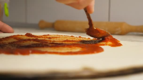 Mužská ruka kuchaře nanáší kečup na těsto na pizzu pomocí lžíce u kuchyňského stolu. Ruka kuchaře namazávající rajčatovou omáčku na pečivo v kovové podobě na dřevěném povrchu v kuchyni. Koncepce přípravy jídla — Stock video