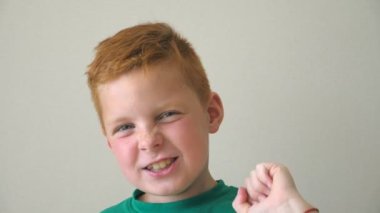 Sevimli yakışıklı çocuk kameraya bakıyor ve sevinçle el kaldırıyor. Yüzünde mutluluk ve sevinç duyguları olan mutlu genç bir çocuk. İçeride çilli bir çocuğun portresi. Ağır çekim. Yaklaş.