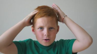 Sevimli kızıl saçlı çocuğun başını tutup yüzünde neşe beliren görüntüsü. Genç bir çocuk duygusal ve duygusal bir şekilde kameraya bakıyor. Kapalı alanda çilli mutlu bir çocuğun portresi. Yavaş çekim