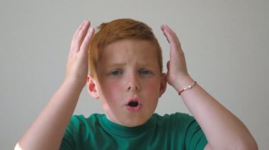 Sevimli kızıl saçlı çocuğun başını tutup yüzünde neşe beliren görüntüsü. Genç bir çocuk duygusal ve duygusal bir şekilde kameraya bakıyor. Kapalı alanda çilli mutlu bir çocuğun portresi. Yavaş çekim