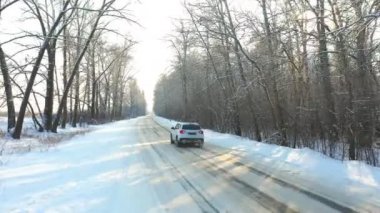 Karla kaplı buzlu yolda giden beyaz arabayı takip ediyorum. Güneşli bir kış gününde ormanda boş kırsal yolda giden bir SUV. Otomobilin manzaralı arazide hareket edişini izlemek. Hava görüntüsü