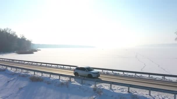 Zdjęcie białego samochodu jadącego przez pokrytą śniegiem drogę w pobliżu zamarzniętego jeziora. SUV jazdy na trasie wsi w pobliżu lodowatego stawu w słoneczny zimowy dzień. Latanie nad samochodem poruszającym się po malowniczym krajobrazie — Wideo stockowe