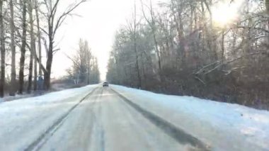 Karla kaplı buzlu yolda giden beyaz arabayı takip ediyorum. Güneşli bir kış gününde ormanda boş kırsal yolda giden bir SUV. Otomobilin manzaralı arazide hareket edişini izlemek. Hava görüntüsü