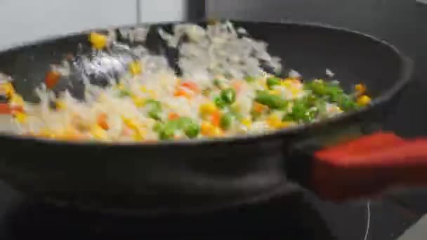 Männliche Köchin hält Pfanne und wirft Reis mit gemischtem Gemüse in die Küche. Chef kocht Risotto mit Mais, Erbsen, Spargel, Paprika und Karotten auf Pfanne. Konzept der Essenszubereitung. Zeitlupe — Stockvideo