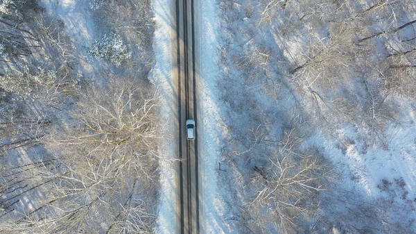 Aerial shot of white car start riding through snow covered icy road. SUV driving at countryside route in the forest on winter day. Flying over the auto moving through scenic landscape way. Top view.