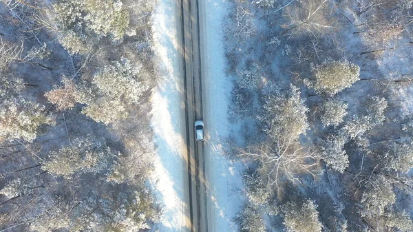 SUV driving at beautiful countryside route in the forest on winter day. Aerial shot of white car ride through snow covered icy road. Flying over the auto moving through scenic landscape way. Top view.