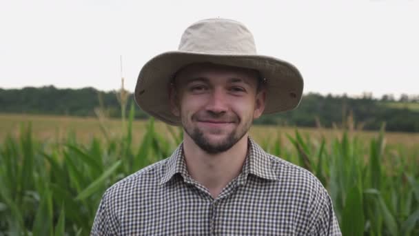 Крупним планом молодий усміхнений фермер в капелюсі дивиться в камеру на розмитому тлі кукурудзяного поля. Портрет щасливого втомленого працівника, що стоїть на кукурудзяному лузі. Концепція аграрного бізнесу — стокове відео