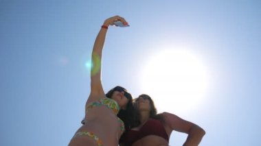 Neşeli, çekici esmerler akıllı telefonda selfie portresi çekiyorlar. Yaz tatilinde fotoğraf çeken kaygısız genç kadınlar. Kameraya poz veren güneş gözlüklü mutlu bayanlar. Kız arkadaşlar birlikte dinleniyor.