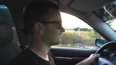 Gözlüklü, mutlu ve yakışıklı bir adamın ülke boyunca araba sürüşünün profili. Otomatik kontrol sırasında yola bakan gülümseyen genç adam. Seyahat konsepti. Arka planda kırsal kesimde gün batımı. Yan görünüm