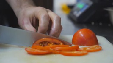 Aşçının erkek elleri mutfak restoranında plastik kesme tahtasıyla taze kırmızı domates kesiyor. Sağlıklı sebze salatası hazırlama süreci. Yemek pişirme kavramı. Yavaşça kapat.