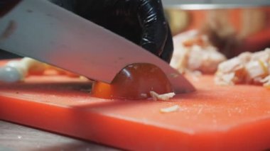 Eldivenli erkek aşçı elleri mutfak restoranında bıçakla plastik doğrama tahtasıyla taze kırmızı domates kesiyor. Sağlıklı sebze salatası hazırlama süreci. Yemek pişirme kavramı. Kapat.