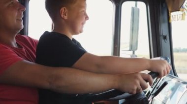 Gülümseyen kızıl çocuk, babasının kucağında oturan büyük direksiyonu tutuyor ve kamyonu sürmeyi öğreniyor. Mutlu baba kızıl saçlı çocuğuna boş yolda araba kullanmayı öğretiyor.