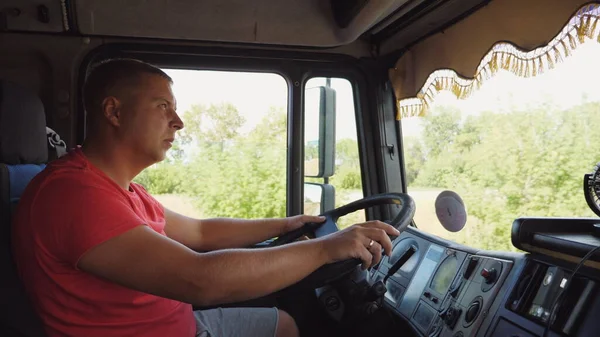 Lorry driver holds arms on a steering wheel and controls the truck riding to destination. Trucker operating a car through countryside. Man driving at country road. Logistics and transportation concept.
