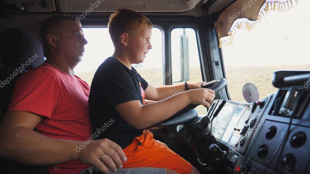 Father teaching his small red-haired kid to operate a car on empty country road. Focused little boy sitting on lap of dad and learning to drive the truck. Dad and son spending time together. Close up.