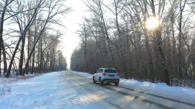 Karla kaplı buzlu yolda giden beyaz arabayı takip ediyorum. Güneşli bir kış gününde ormanda boş kırsal yolda giden bir SUV. Otomobilin manzaralı arazide hareket edişini izlemek. Hava görüntüsü