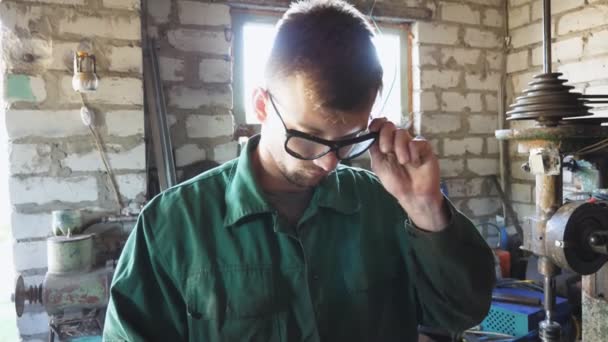 防護アイウェアを脱ぎ、額から汗を拭く作業着の男性整備士。若い修理工が眼鏡をかけガレージで仕事を続ける。保守サービスの概念。閉じろ! — ストック動画