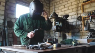 Koruyucu maskeli genç bir tamirci bozuk kaynak makinesi tutuyor ve garajda bazı metal parçaları lehimlemeye çalışıyor. Atölyesinde çalışan erkek kaynakçı. Bakım hizmeti kavramı. Kapat.