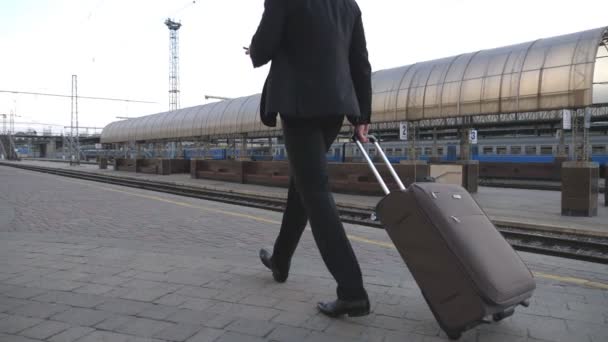 Homme d'affaires prospère méconnaissable marchant à travers la gare et tirant une valise sur roues. Un homme d'affaires avec ses bagages se promenant près du chemin de fer et utilisant un smartphone. Ralenti Fermer — Video