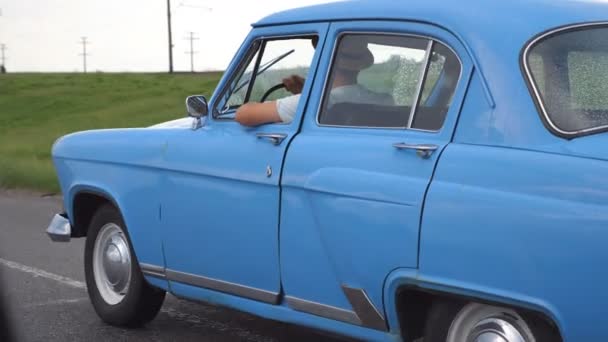 Młoda para w kapeluszach jadąca niebieskim samochodem retro po deszczu podczas letnich podróży. Nierozpoznawalna para poruszająca się starym, zabytkowym pojazdem na autostradzie. Zwolniony ruch Zamknij widok z boku — Wideo stockowe