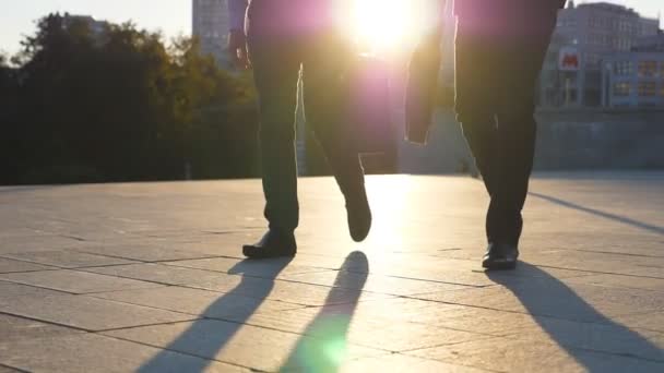 Füße von zwei Geschäftsleuten mit Aktentaschen, die bei Sonnenuntergang auf der Straße spazieren gehen. Partner pendeln zur Arbeit. Selbstbewusste Typen auf dem Weg ins Amt. Kollegen, die nach draußen gehen. Zeitlupe — Stockvideo