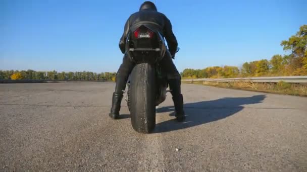 Achteraanzicht van onherkenbare motorrijder in helm en leren jas zit op de motorfiets op de snelweg en klaar om te rijden. De man gaat op een motor rijden. Concept van vrijheid en avontuur. Sluiten. — Stockvideo