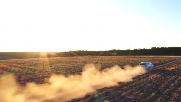 Tiro aéreo de um carro eléctrico a mover-se pela estrada, deixando rasto de poeira para trás. Ecologia auto amigável em carga elétrica dirigindo ao longo da estrada rural. Paisagem de pôr do sol cênica no fundo — Vídeo de Stock