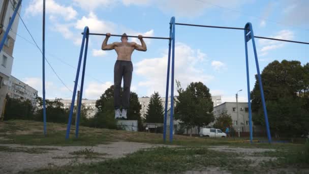 Atleta muscular haciendo flexiones en el campo de deportes. Joven deportista ejercitándose en barra horizontal en el patio de recreo. Hombre deportivo fuerte entrenamiento al aire libre. Concepto de estilo de vida deportivo. Dolly disparó de cerca — Vídeos de Stock