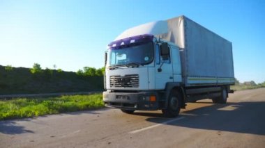 Kamera, kargo karavanıyla otoyolda giden ve malları taşıyan kamyonu takip ediyor. Güneşli bir günde kırsal yolda hızla giden bir kamyon. Güneş arka planda parlıyor. Yavaş çekim. Yakın çekim.