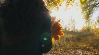 Elinde bir buket sarı akçaağaç yaprağı ile sonbahar ormanında yürüyen tanınmayan bir kadın. Hafif esintinin ve güzel doğal manzaranın tadını çıkaran bir kız. Güneş arka planda parlıyor. Yavaş çekim. Yakın çekim.