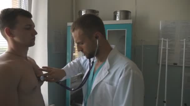 De dokter checkt de borst van een man in haar kantoor in het ziekenhuis. Dokter onderzoekt jonge mannelijke patiënt met stethoscoop. Medisch werker luisterend naar de hartslag van atletische man. Sluiten. — Stockvideo