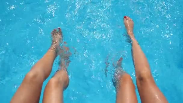 Vista superior sobre pernas femininas salpicando água cristalina na piscina do hotel. Dois amigos sentados à beira da bacia e desfrutando de suas férias no dia ensolarado. Conceito de férias de verão ou férias. Devagar. — Vídeo de Stock