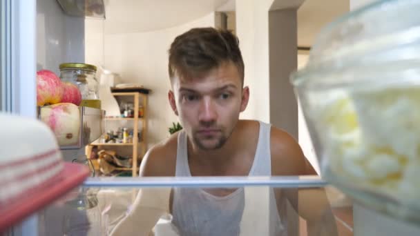 Handsome young guy opens kitchen refrigerator looking inside suspecting that there is something or somebody. Confused man making sure that there is nothing but food in fridge and closing it. POV — Stock Video