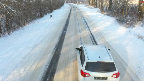 Biały samochód szybko przejeżdża przez pokrytą śniegiem lodową drogę. SUV jedzie na pustą wiejską trasę w zimowym lesie w słoneczny dzień. Auto poruszające się po malowniczym krajobrazie. Koncepcja podróży. Widok z góry Drone shot — Wideo stockowe