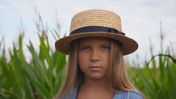 Portrait of small serious girl in straw hat looking into camera against the blurred background of corn field at organic farm. Little kid with long blonde hair standing in the meadow. Close up — Stock Video