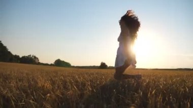 Beyaz elbiseli tanınmayan bir kadın gün batımında sarı buğdayla tarlada koşuyor. Genç kaygısız kız güzel doğa ortamında özgürlüğün tadını çıkarıyor. Manzaralı yaz manzarası. Yan Görünüm Kapat