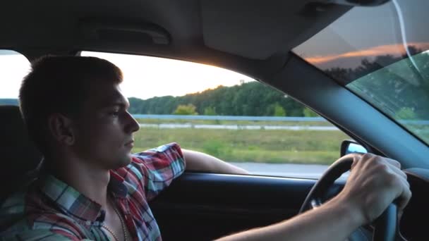 Profil przystojnego mężczyzny w koszuli jeżdżącego samochodem przez wieś. Młody człowiek jadący samochodem z ręką za oknem, cieszący się podróżą. Piękny widok na tle. Powolny ruch Zamknij — Wideo stockowe