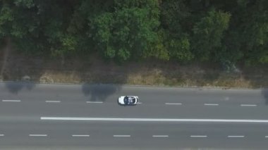 Aerial view of white convertible car riding along country road. Young couple traveling on cabriolet. Happy just married pair enjoying trip together. Flying over auto driving on highway at summer day