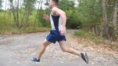 Kablosuz kulaklıklar ile yakışıklı güçlü sporcu orman yakınındaki iz boyunca hızlı sprint. Doğada yol boyunca koşan sportif bir adam. Atletik adam erken sonbaharda egzersiz. Sağlıklı aktif yaşam tarzı