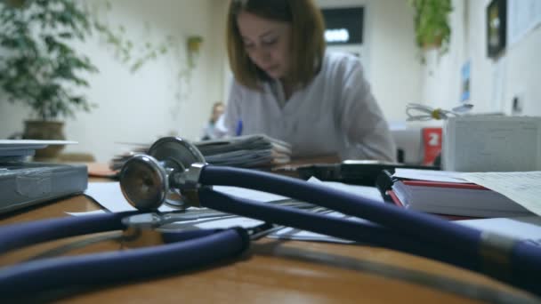 Młoda pielęgniarka pracująca w biurze, pisząca dokumentację medyczną, stetoskop na pierwszym planie. Kobieta-lekarz wypisuje receptę w szpitalu. Koncepcja opieki zdrowotnej. Zamknij się. — Wideo stockowe