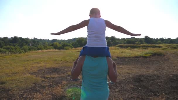 Far och son leker på sommarplanen på solig dag. En liten pojke som sitter på sin pappas axlar och spelar höjda händer som ett flygplan. Förälder med barn som har roligt tillsammans. Slow motion Bakifrån — Stockvideo