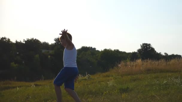 Mladý chlapec se zdviženýma rukama pobíhající po zelené trávě na louce za slunečného dne. Děti běhají venku na trávníku. Šťastně se usmívající chlapec baví v přírodě na letní louce. Uzavření pomalého pohybu — Stock video