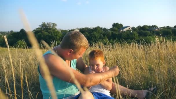 Glad ung man som ligger på gräset och leker med sin lilla pojke. Far kittlar sin son utomhus. Glada pappa och hans barn har kul tillsammans i en park. Långsam rörelse Närbild — Stockvideo