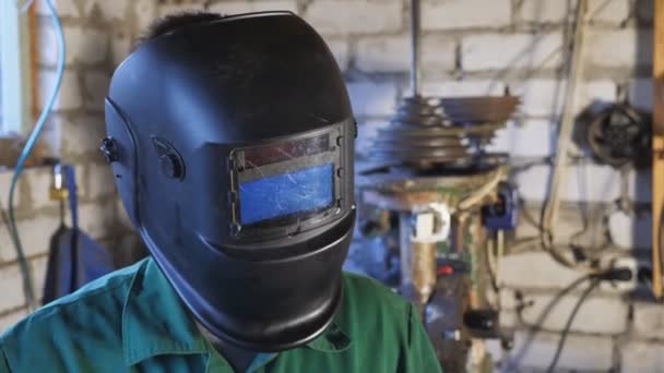 Jonge reparateur in uniform opent een beschermend masker na zijn werk. Vermoeide lasser kijkt in de camera in zijn garage. Portret van monteur met baard aan het werk in de werkplaats. close-up Dolly schot — Stockvideo