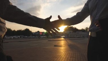 İki işadamı, arka planda güneş ışığıyla şehir ortamında buluşup birbirlerini selamlıyorlar. Dışarıda iş tokalaşması. Erkek kollarını dışarıda sallıyor. Meslektaşlar el sıkışır Yavaş çekim kapanır