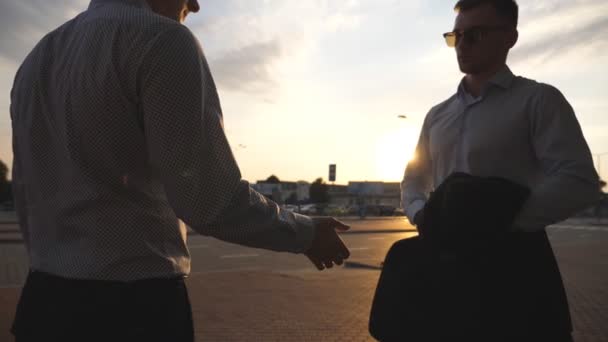 Business man passing a black briefcase to his partner. Colleagues shake hands outdoor in the city background. Two young businessmen meeting outdoor and greeting each other. Close up Slow motion — Stock Video