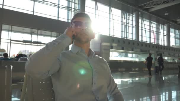 Jeune homme d'affaires parlant au téléphone à l'aéroport avec éruption de soleil en arrière-plan. Gros plan portrait d'un bel homme d'affaires en lunettes de soleil parlant sur son téléphone portable dans le hall du bureau. Mouvement lent — Video