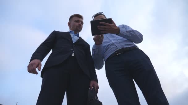 Low angle view of young businessman showing a presentation on screen of tablet pc to his colleague in city. Successful entrepreneurs working on digital tablet. Blue sky at background. Slow motion — Stock Video