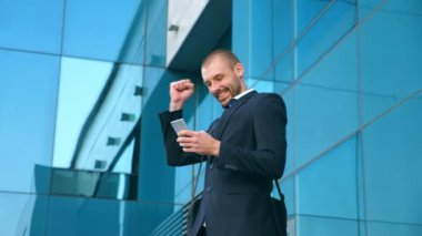 Genç işadamı ofis yakınında akıllı telefon kullanıyor ve başarıyı kutluyor. İş adamı telefonda iyi haberleri ve olumlu duyguları öğrendi. Erkek portresi başarıyla mutludur. Yavaş çekim